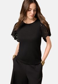 Black t-shirt with scalloped lace sleeves and a round neckline. Smooth fabric with a fitted design, paired with loose black trousers.