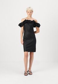 Μαύρο φόρεμα με ανοιχτούς ώμους και βολάν μανίκια, φτιαγμένο από γυαλιστερό ύφασμα. Εφαρμοστό σιλουέτα, με μήκος μέχρι το γόνατο, σε συνδυασμό με μαύρες σαγιονάρες.