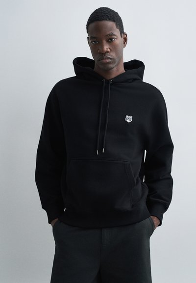 Schwarzer Kapuzenpullover aus strukturiertem Stoff, mit einem kleinen gestickten Wolf-Logo auf der Brust und einer Fronttasche.