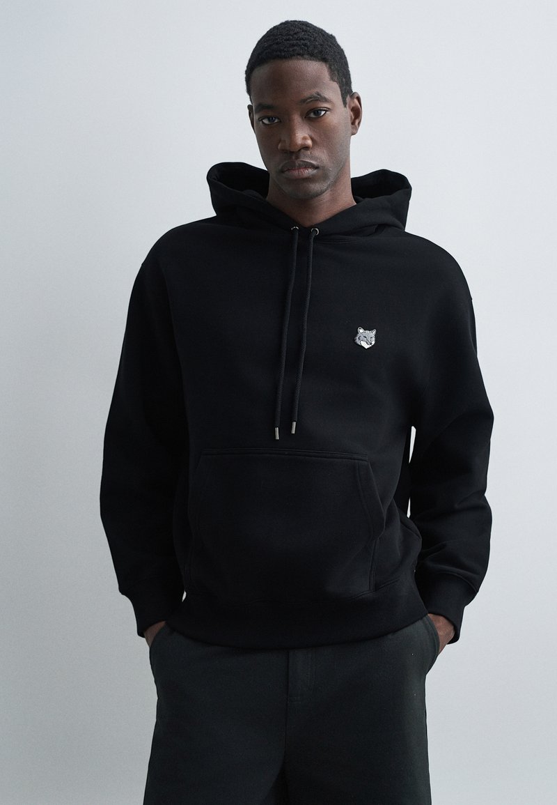 Sweatshirt à capuche noir en tissu texturé, présentant un petit logo de loup brodé sur la poitrine et une poche frontale.