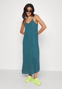 Robe maxi turquoise avec un motif blanc subtil dispersé, bretelles spaghetti et un ourlet évasé. Associée à des crocs jaune vif.
