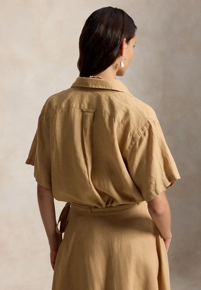 Tamsių plaukų moteris, dėvinti šviesios spalvos trumpomis rankovėmis lininę palaidinę ir sijoną, matoma iš nugaros neutraliame fone.