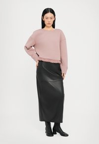 Sweatshirt scurt de culoare roz deschis, cu un tiv ondulat, asociat cu o fustă maxi din piele neagră. Modelul poartă cizme negre până la gleznă. Fundal simplu.