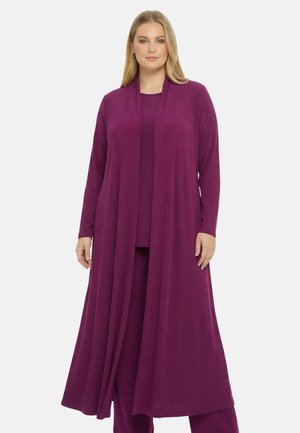 Vrouw draagt een lange, geribbelde, paarse cardigan over een bijpassende paarse top en broek, staand voor een effen witte achtergrond.