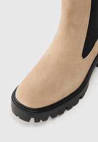 Beige semsket ankelstøvler med en kraftig sort gummisål, som har elastiske sidepaneler for enkel påkleding og en strømlinjeformet tådesign.