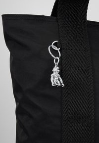 Zwarte stoffen draagtas met een brede band, voorzien van een zilveren sleutelhanger met een zittend figuur. De textuur is glad met een lichte glans.