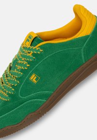 Zapatilla de ante verde con acentos amarillos, cordones texturizados y suela de goma marrón. Presenta una forma redondeada y una lengüeta acolchada.