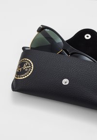 Ochelari de soare Ray-Ban într-un toc din piele neagră cu o suprafață texturată, având un logo auriu și un închidere cu capse argintie.