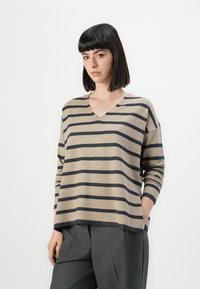 Kvinne med rett, mørkt hår som har på seg en beige og svart stripete V-halsgenser og grå bukser, poserer med den ene hånden i lommen.