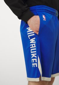 Pantalones cortos de baloncesto azules con tejido de malla transpirable, aberturas laterales y texto bordado "MILWAUKEE" en blanco en la pierna izquierda.