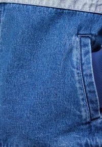Close-up of blue denim jeans pocket with visible stitching and texture, worn with a denim jacket and blue shirt.