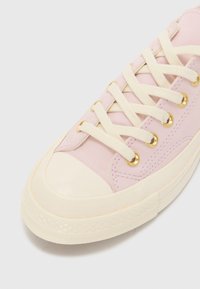 Pink canvas sneaker with gold eyelets, cream laces, and a rubber toe cap. Features textured sole and contrasting stitching.