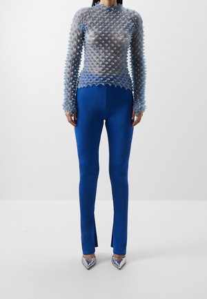 Femme portant un haut transparent à manches longues avec des pointes texturées en 3D, un pantalon ajusté bleu royal avec des fentes à l'avant, et des talons argentés à bouts pointus.