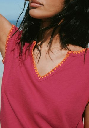 Woman with black hair wearing a magenta top with orange zigzag trim around the V-neck and sleeves, shown from shoulders to chin.