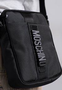 Svart nylon crossover-väska med rektangulär form, utrustad med upphöjd "MOSCHINO"-logotyp, texturerad yta och justerbar axelrem.