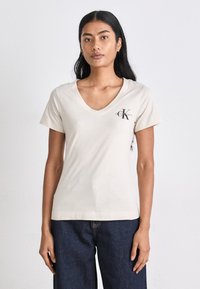 Šviesiai smėlinė v-neck marškinėliai iš minkštos medvilnės, trumpomis rankovėmis ir logotipu kairėje krūtinės pusėje. Su tamsiai mėlynomis džinsomis.