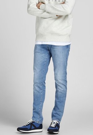 Person stående med armene krydsede iført en lysegrå sweatshirt, lyseblå jeans og blå og sorte sneakers.
