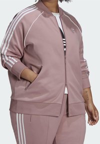 Chaqueta bomber deportiva rosa con rayas blancas en las mangas, cierre de cremallera y puños acanalados. Cuenta con bolsillos laterales y textura lisa.
