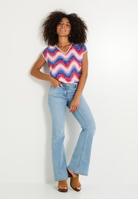 Top colorido con patrón ondulado en rosa, rojo y azul; combinado con jeans acampanados de un tono azul claro y sandalias de tacón color beige. Presenta un diseño de escote en V.