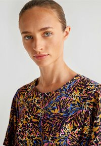 T-shirt w multikolorowym, żywym, abstrakcyjnym wzorze w odcieniach fioletu, niebieskiego, złota i czerni. Miękka tkanina, okrągły dekolt, krótkie rękawy.