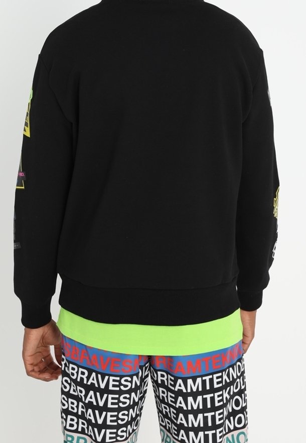 Personne portant un sweat-shirt noir et un short coloré avec des motifs de texte répétés "BRAVES" et "DREAMTEK", debout de dos.