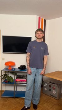 Νεαρός άνδρας με τατουάζ στα χέρια στέκεται σε δωμάτιο με τηλεόραση στον τοίχο, πορτοκαλί φωτιστικό, κάμερα, βιβλία, φυτό και ασημένια αυτοκόλλητα βαλίτσας στο ξύλινο πάτωμα.