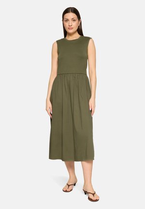 Vrouw staand met een mouwloze olijfgroene midijurk met geplooide taille en bruine sandalen met lage hak op een effen achtergrond.