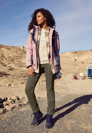 Light pink jacket over a plaid shirt, olive green pants, and dark hiking boots. Outdoor setting with rocky terrain and blue sky.