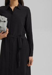 Czarna sukienka z długim rękawem, z kołnierzykiem zapinanym na guziki, paskiem w talii oraz rękawami z mankietami. Modelka trzyma szary torebkę w kształcie pudełka.