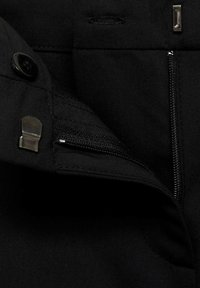 Țesătura neagră cu o textură netedă are o buzunar cu fermoar și accesorii metalice. Include o închidere cu buton și detalii de cusătură subtile.