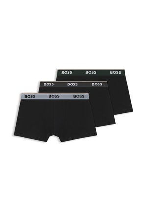 Muskelbepackter Mann steht mit den Händen in der Hüfte, trägt schwarze Boxershorts mit der Aufschrift BOSS am Bund vor schlichtem weißem Hintergrund.