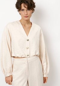 Crème cropped blouse met een V-hals, textuurstof, ballonmouwen en zilveren knopen. Gecombineerd met lichtgekleurde bijpassende broeken.