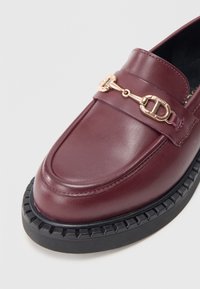 Burgundy läderloafer med rundad tå, med en kedjedetalj i guldton och en texturerad svart gummisula.