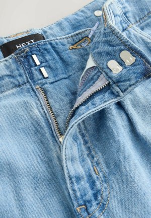 Jean en denim bleu clair avec braguette zippée, fermeture à bouton en métal et surpiqûres contrastantes. Étiquette visible à la taille.