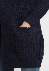 Námořnický pletený kardigan s žebrovanou texturou, dvěma předními kapsami a dlouhými rukávy. Měkká příze poskytuje pohodový vzhled.