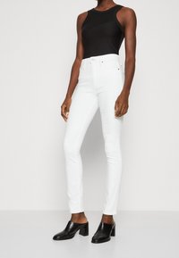 Jeans skinny de tiro alto blancos en tejido elástico, combinados con un body sin mangas negro y mulas negras con tacón, que muestran una silueta elegante.