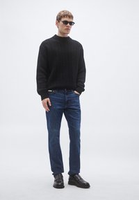 Suéter negro tejido con cuello alto, jeans azules y zapatos de cuero negros. La modelo lleva gafas de sol oscuras frente a un fondo neutro.