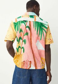 Camisa de manga corta con un diseño en amarillo claro y rosa, que presenta patrones tropicales, hojas de palma verdes y formas abstractas.