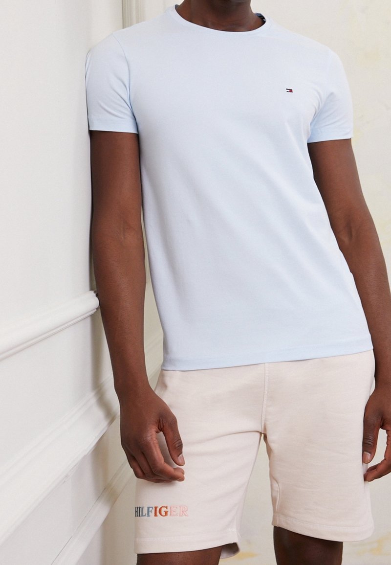 Hellblaues kurzärmliges T-Shirt mit Rundhalsausschnitt, getragen mit beigen Shorts, die das Logo "HILFIGER" in kontrastierenden Farben zeigen.