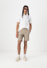 White polo shirt with blue logo, tan shorts, light sneakers, and sunglasses. Straight fit, casual style, minimal detailing.