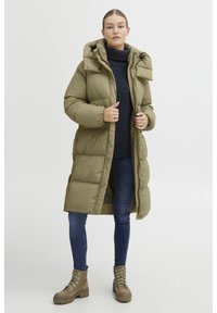 Manteau long matelassé vert olive avec une capuche, un col montant, des boutons-pression et des poches latérales, associé à un jean foncé et des bottes en cuir à lacets marron.