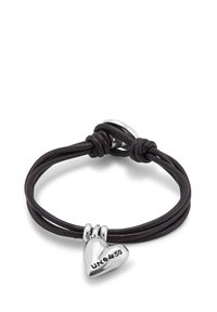 Pulsera de cuero en marrón oscuro con un encanto en forma de corazón de plata; presenta una superficie texturizada y un logotipo grabado en el encanto. Cierre ajustable.