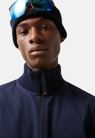 Navy blauwe zip-up jack met geribde kraag en ritsdetails. Draagt een zwarte beanie en zonnebril met blauwe reflecterende glazen.