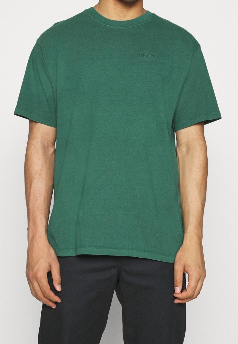 Homme portant un t-shirt uni vert foncé à manches courtes et col rond, avec un pantalon noir, debout devant un fond blanc.