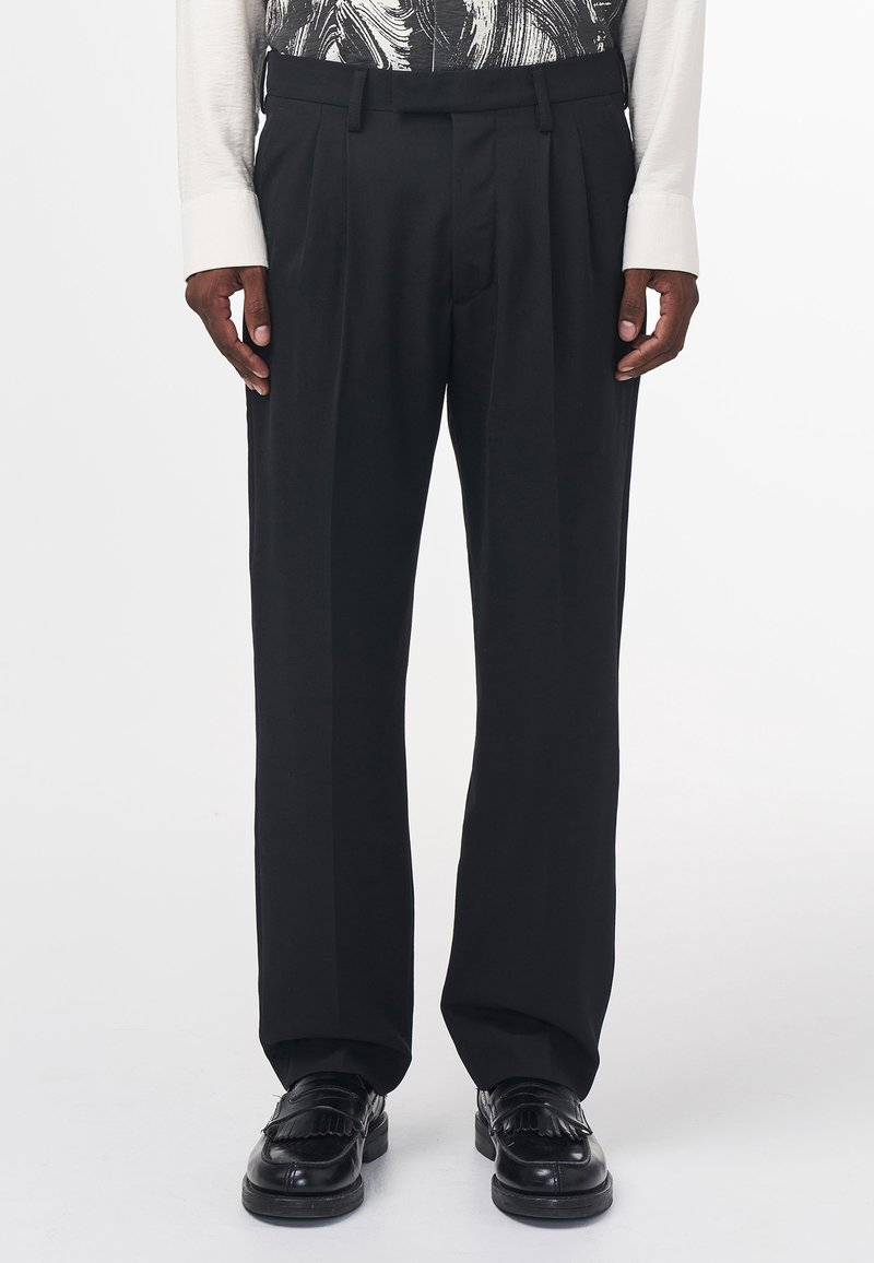 Homme portant un pantalon noir plissé, ample, avec poches latérales et des mocassins en cuir noir, debout devant un fond blanc uni.