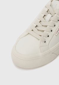 Primo piano di una sneaker beige Calvin Klein con suola testurizzata e lacci in tessuto su uno sfondo bianco uniforme.