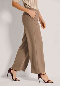 Vrouw in beige wijde broek en bordeauxrode hoge hakken, wandelend met één hand in de zak tegen een effen achtergrond.