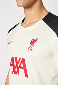 Maglietta del Liverpool FC, color crema con spalle nere, presenta logo e scritte rossi. Realizzata in tessuto testurizzato, con scollatura rotonda.