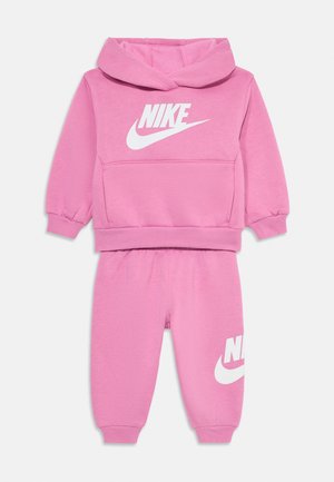 Roza Nike trenirka, ki vključuje kapuco z belim logotipom, kengurov žep in usklajene hlače z elastiko na robovih ter logotipom na nogi.