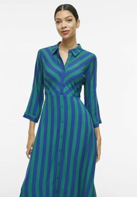Dolga srajčna obleka z gumbi, okrašena z navpičnimi modrimi in zelenimi črtami. Tkanina se zdi gladka, s prilagojenim krojem in ovratnikom.